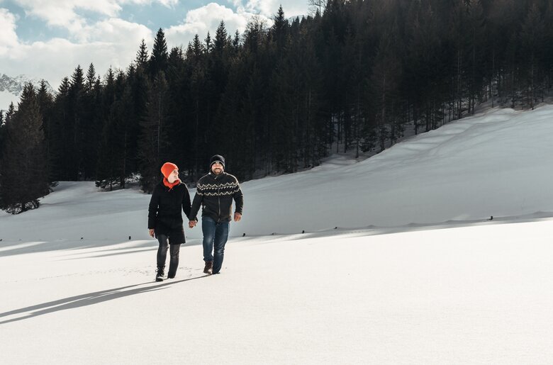 Two people walking together in a snowy winter landscape with a forested mountainous background. | © Urlaub am Bauernhof / Punkt und Komma