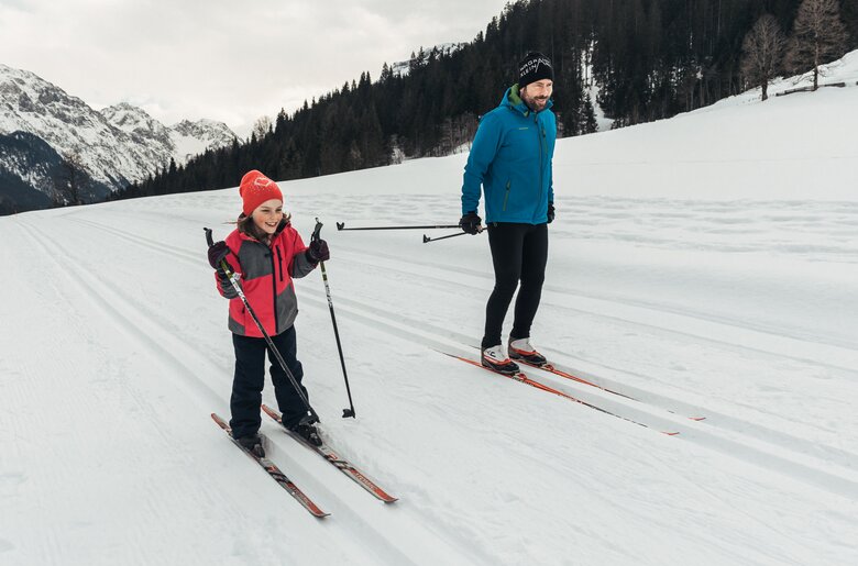 Langlaufen | © Urlaub am Bauernhof / Punkt und Komma Two people, a woman in red winter gear and a man in blue, skiing on a snowy mountainous landscape. | © Urlaub am Bauernhof / Punkt und Komma