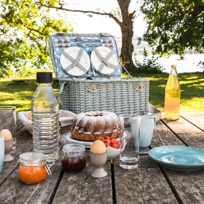 Picnic basket, cake, strawberries, drinks, and tableware on wooden table in garden with lush greenery. | © Urlaub am Bauernhof / Punkt und Komma