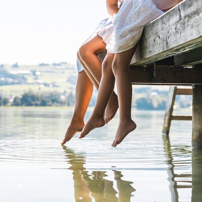 Person's legs dangling over a wooden dock, reflecting on the calm water surface, with a scenic landscape in the background. | © Urlaub am Bauernhof / Punkt und Komma