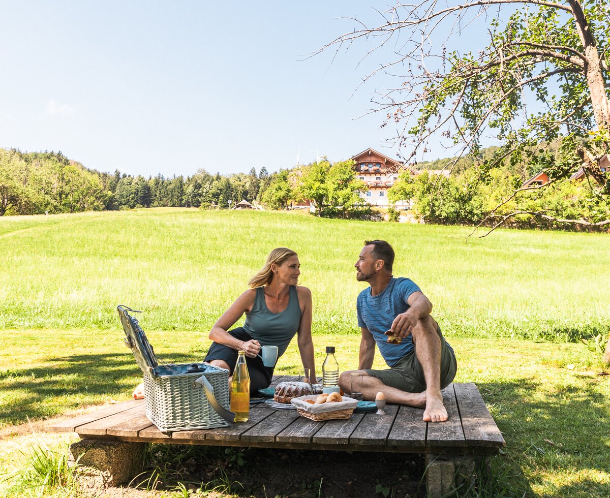 Couple on picnic blanket in meadow, enjoying lunch and each other's company, with a cabin in the background. | © Urlaub am Bauernhof / Punkt und Komma