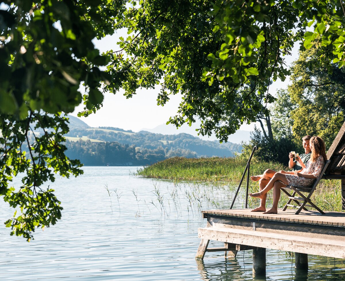 Lakeside cabin, woman relaxing on wooden dock surrounded by lush greenery, tranquil lake landscape in the background. | © Urlaub am Bauernhof / Punkt und Komma