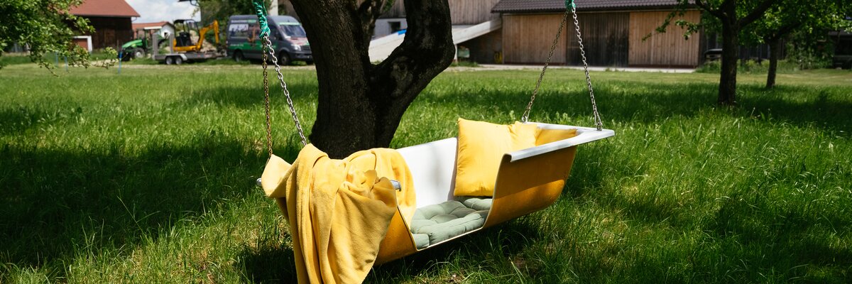 A yellow swing hanging from a tree in a grassy field, with a wooden farmhouse visible in the background. | © Daniel Gollner / Urlaub am Bauernhof
