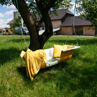 A yellow swing hanging from a tree in a grassy field, with a wooden farmhouse visible in the background. | © Daniel Gollner / Urlaub am Bauernhof