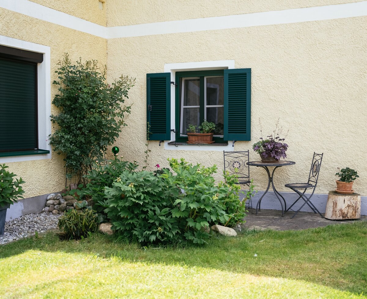 Well-maintained garden with lush greenery, potted flowers, and small patio furniture in front of a cozy, yellow-painted house with green shutters. | © Daniel Gollner / Urlaub am Bauernhof
