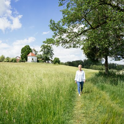 Grassy field, path winding through trees, small chapel on hilltop, woman walking in casual attire, serene countryside landscape. | © Daniel Gollner / Urlaub am Bauernhof