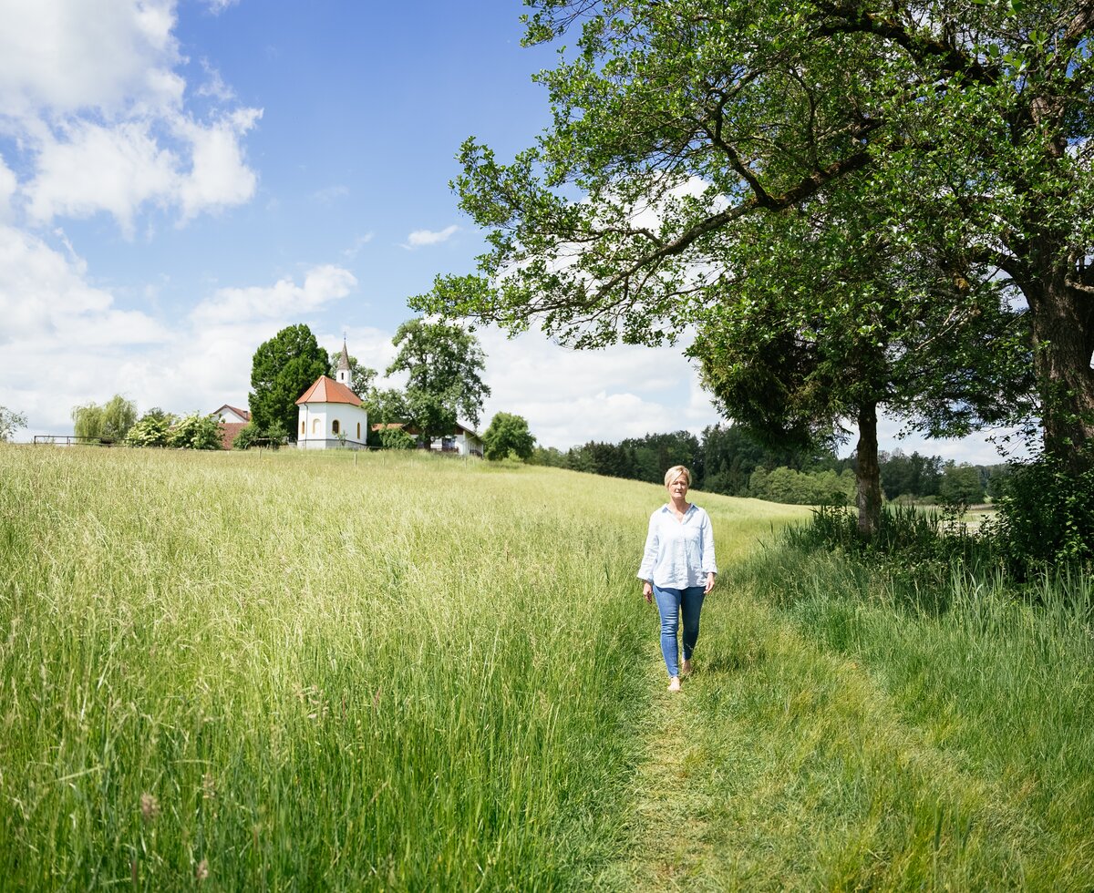 Grassy field, path winding through trees, small chapel on hilltop, woman walking in casual attire, serene countryside landscape. | © Daniel Gollner / Urlaub am Bauernhof