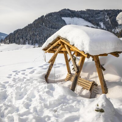 eingeschneite Schaukel | © Urlaub am Bauernhof / Bernd Suppan Snow-covered wooden shelter with benches in a picturesque winter landscape with mountains and forests. | © Urlaub am Bauernhof / Bernd Suppan