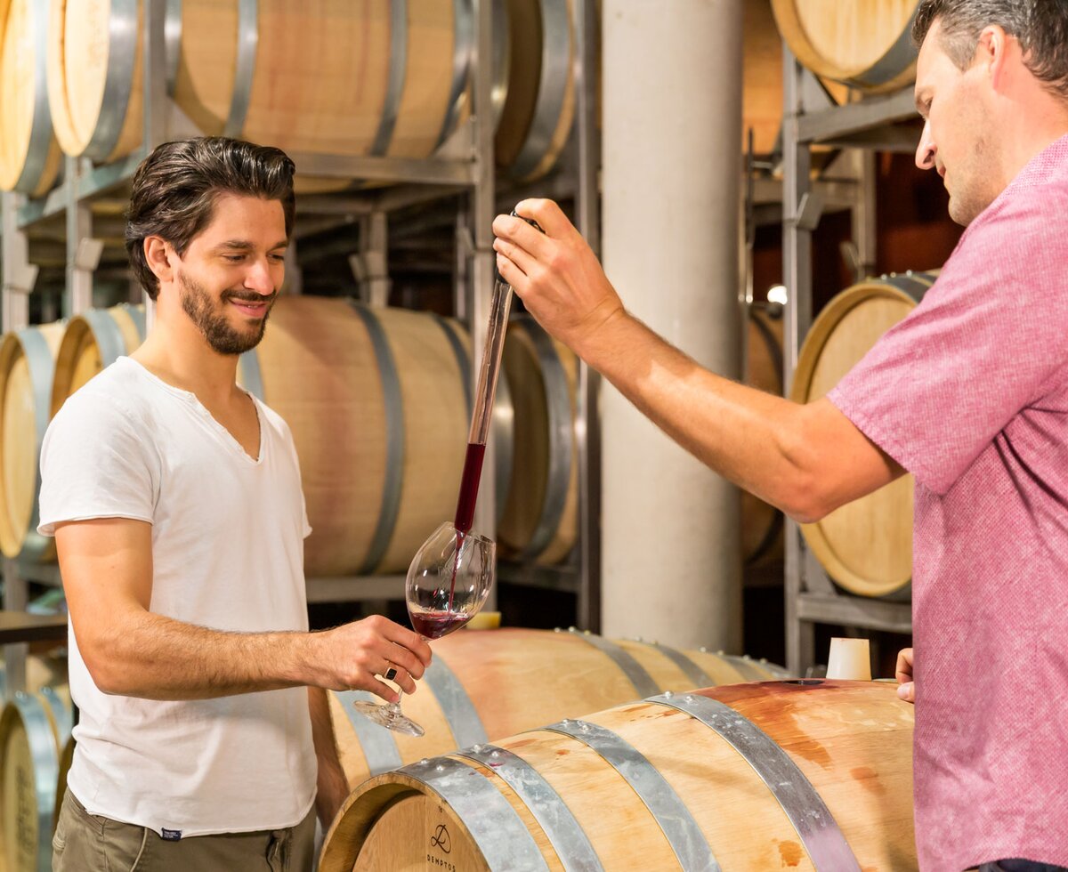Two men tasting wine in a winery, surrounded by wooden barrels.