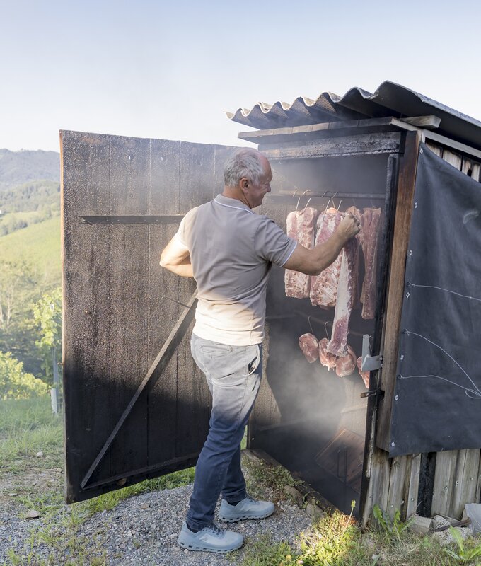 An elderly man tending to hanging meat in a rustic wooden structure, with a scenic countryside view in the background.