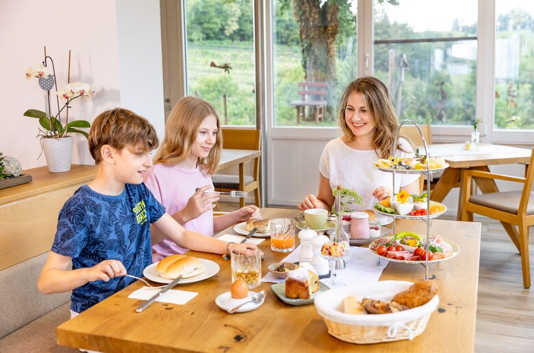 Family enjoying a homemade afternoon tea with pastries, fruit, and beverages on a wooden table by a window overlooking a peaceful outdoor scene.