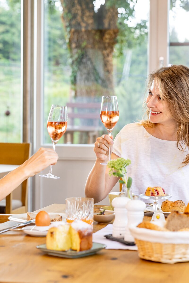 Two women enjoying a tea party with desserts and beverages in a cozy home setting with a scenic outdoor view.