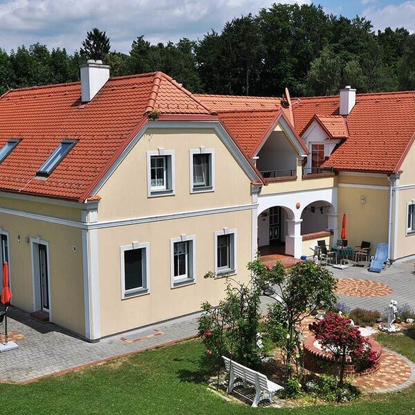 The farmhouse features a light yellow facade, red tiled roof, multiple windows, and exterior terraces with seating areas, surrounded by a garden. The farmhouse features a light yellow facade, red tiled roof, multiple windows, and exterior terraces with seating areas, surrounded by a garden.
