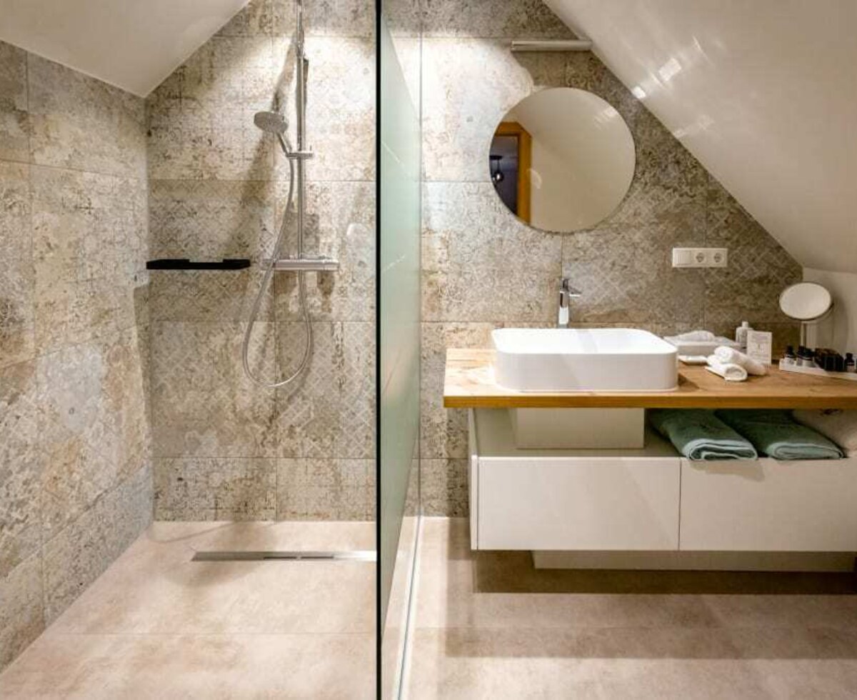 The bathroom in the Farm House includes a glass-enclosed shower, a vanity with a white basin sink, and a round mirror under a sloped ceiling.