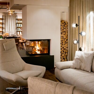 A living area in the farm house includes a sofa, an armchair, a modern glass-fronted fireplace with stacked firewood, and a dining space equipped with bar stools.