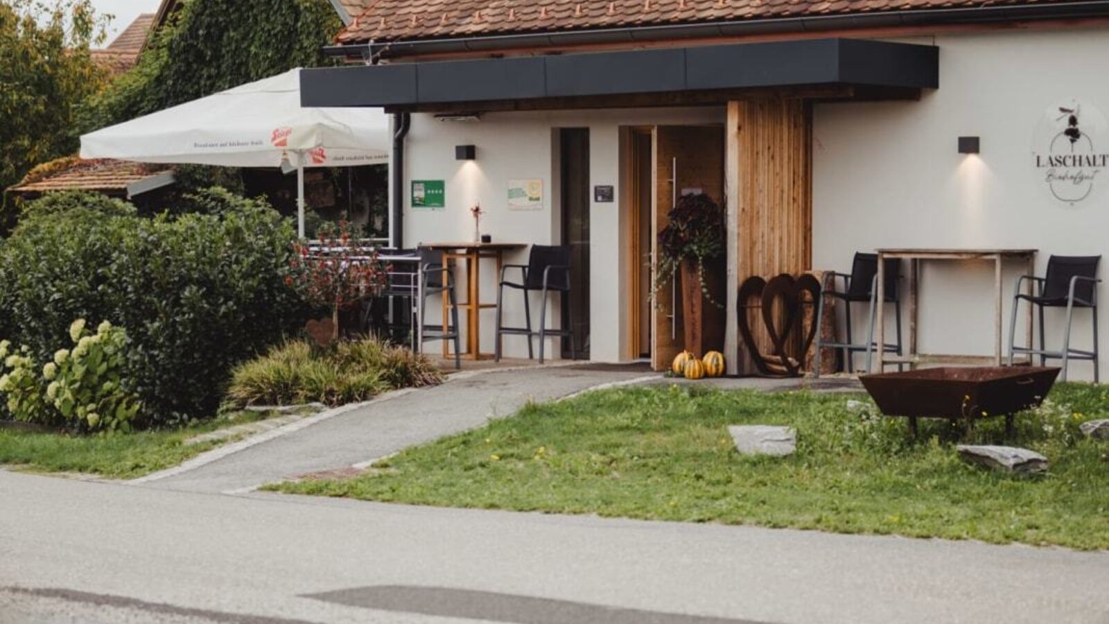 Exterior view of the farmhouse with a white facade, brown tiled roof, and outdoor seating areas; a cat crosses the road in the foreground.