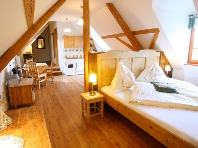 A wooden bed, dining table, and kitchenette area with wooden cabinets and flooring, located in the living space of the farmhouse.