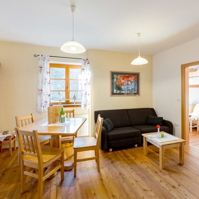 Living area in the farmhouse featuring a dining table, sofa, wooden floor, and an adjacent bedroom with bunk beds.