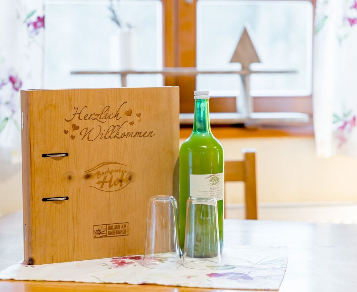 A wooden welcome binder with 'Herzlich Willkommen' (Welcome), a bottle of juice, and glasses on the farm's table.