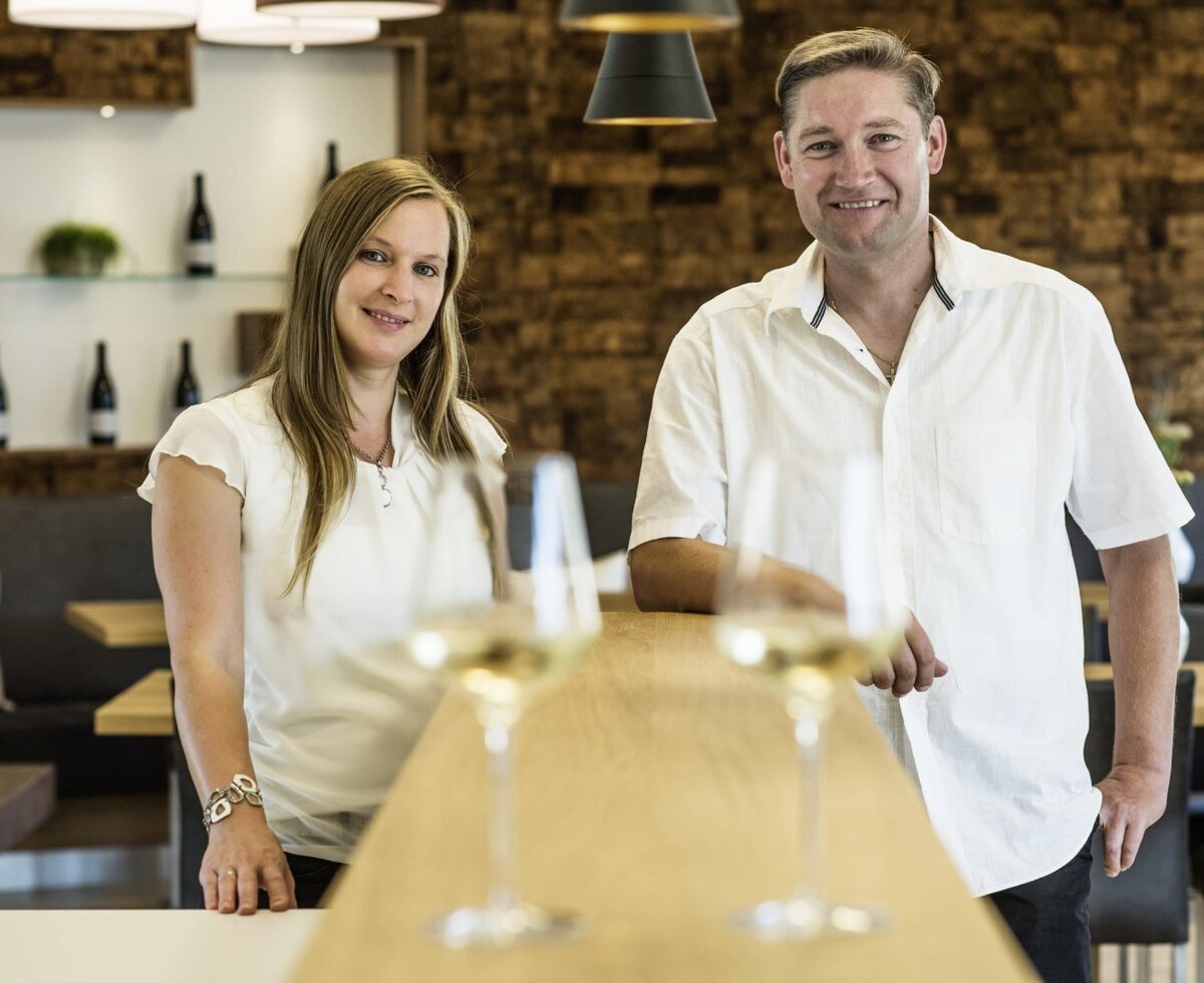 The hosts of the farmhouse welcome guests from behind a counter, with wine glasses and a dining area in the background.