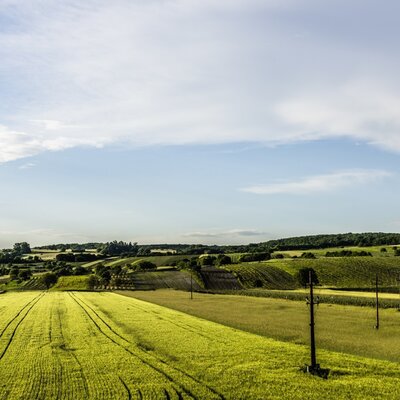 The surrounding landscape of the farmhouse, featuring cultivated green fields and rolling hills under a blue sky.