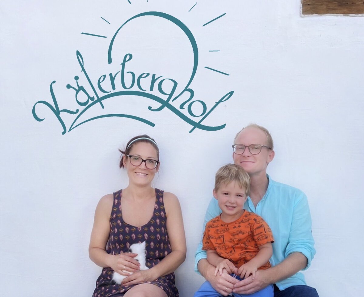 A family with a small white pet sits on a bench at the Farm House, with the Käferberghof logo painted on the wall.