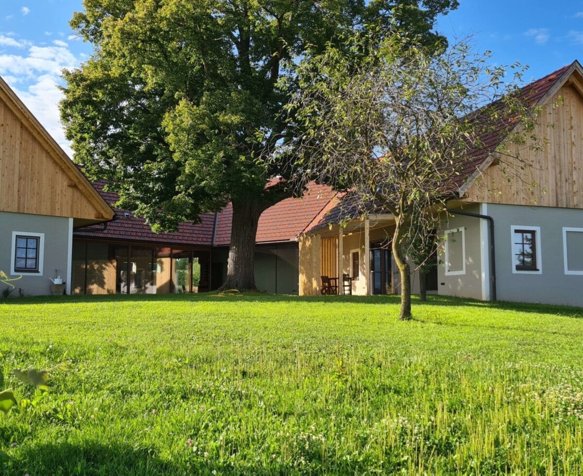 The farmhouse exterior features two buildings with grey walls and wooden gables, a large central tree, a covered patio with outdoor seating, and a spacious surrounding lawn.
