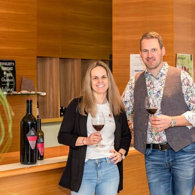 The hosts of the farmhouse, holding wine glasses, stand by a counter displaying wine bottles.
