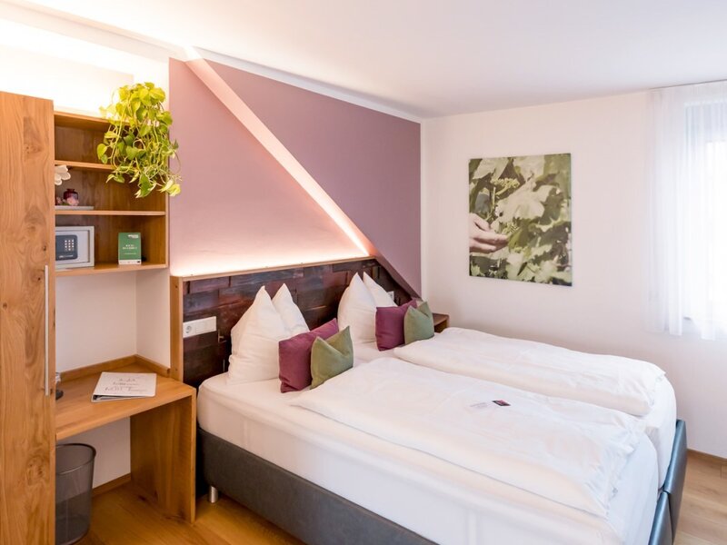 Farmhouse bedroom featuring two single beds, a wooden headboard with ambient backlighting, a built-in desk with shelving and a safe, and light hardwood flooring.