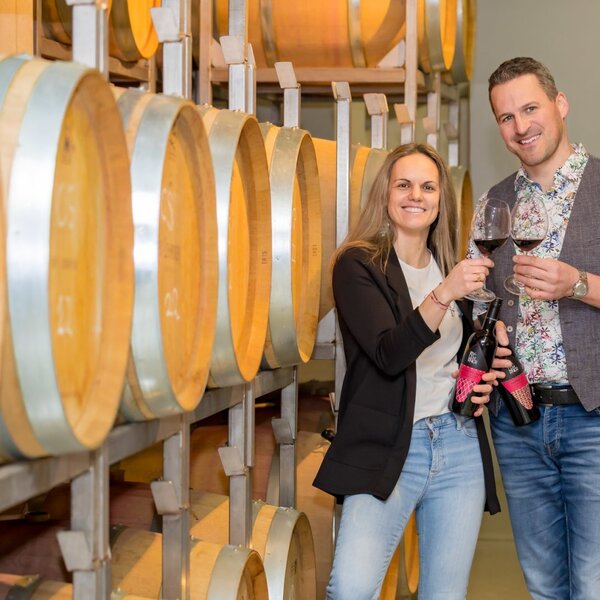 Two individuals hold wine glasses and bottles in the farmhouse's barrel storage room, featuring its own wine.
