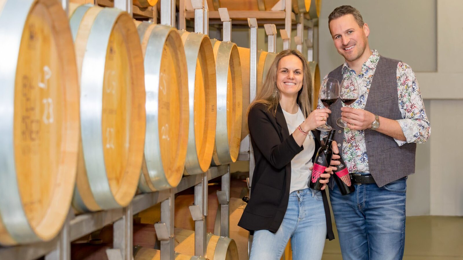 Two individuals hold wine glasses and bottles in the farmhouse's barrel storage room, featuring its own wine.