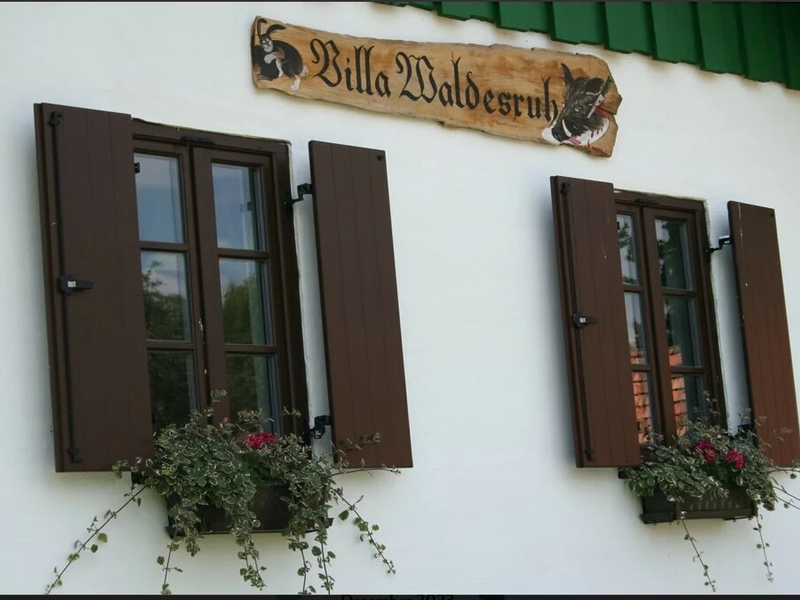 The farmhouse exterior, displaying the "Villa Waldesruh" sign above windows adorned with shutters and floral window boxes.