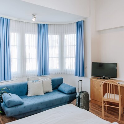 Room in the house with a bed, blue sofa, bay window, cable TV, desk, and wardrobe.