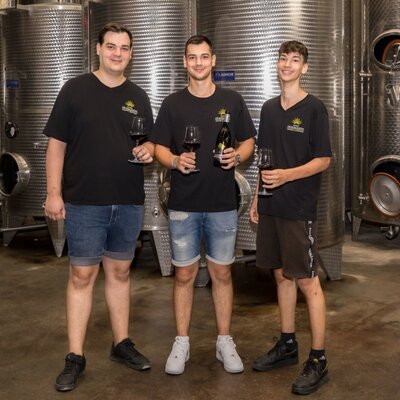Farmhouse staff members in the winemaking facility, holding glasses of wine and a bottle, next to stainless steel fermentation tanks.
