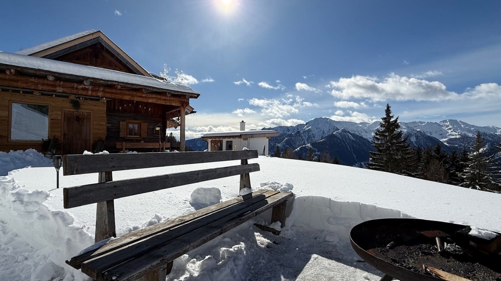 The Alm features a wooden bench and fire pit in the snowy outdoor area, offering mountain views.