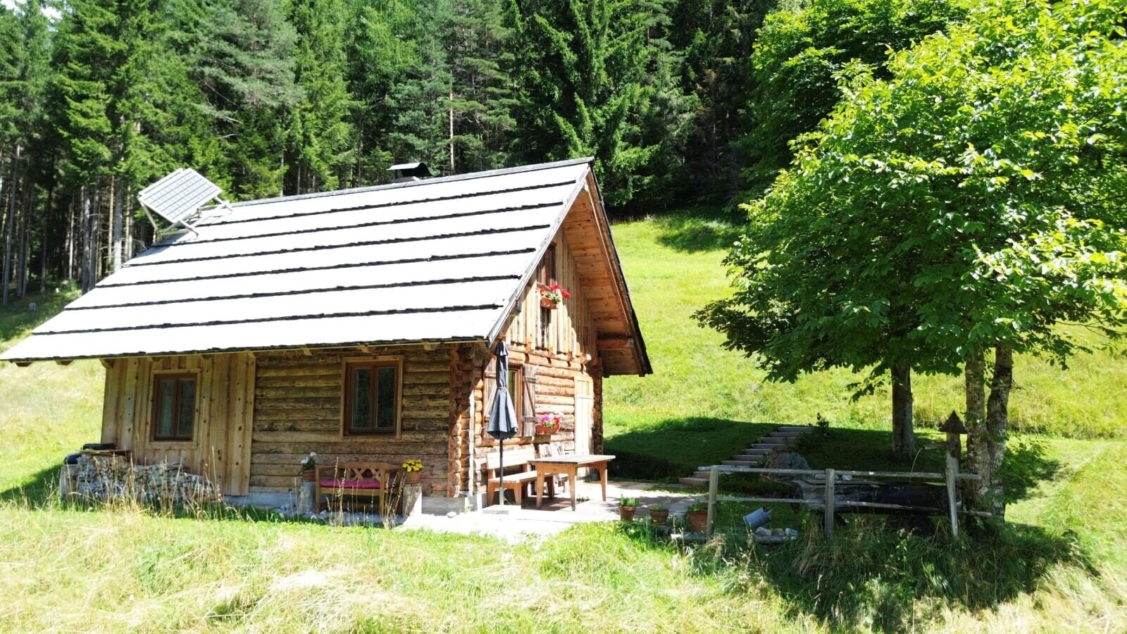 alpine hut with solar panel, and an outdoor seating area, surrounded by meadows and forest.