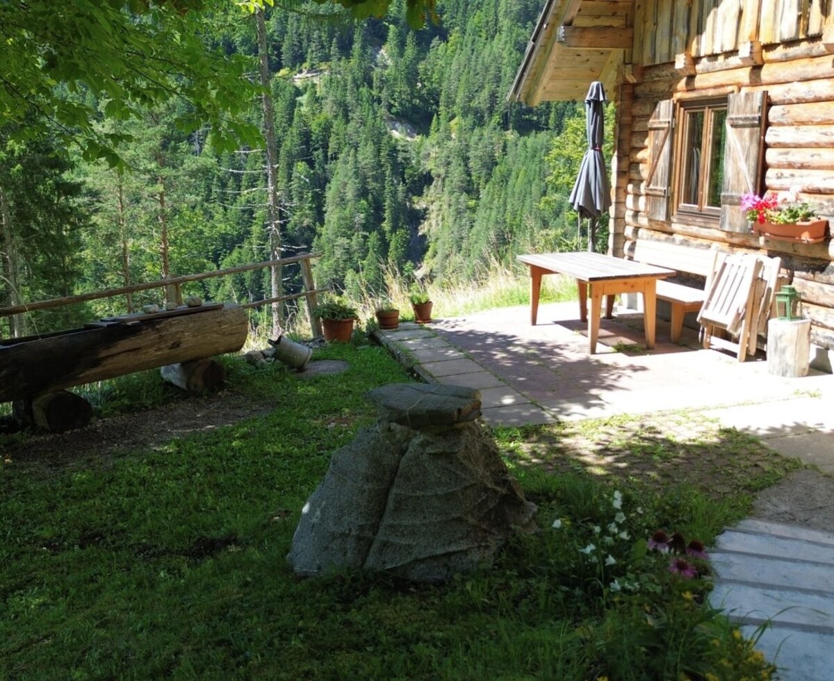 The terrace of the alpine hut with a wooden table and benches offers views of the forested mountains.