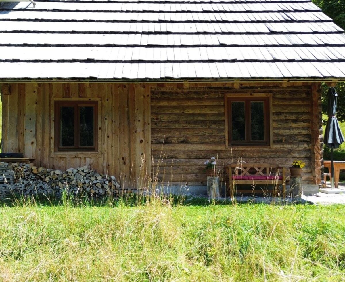 The rustic alpine hut with a shingled roof, two windows, stacked firewood, and a patio with a table and umbrella.