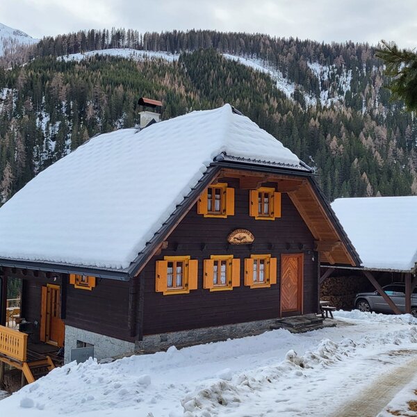 The Alpine Hut exterior, featuring a snow-covered roof, yellow window shutters, a wooden balcony, and a covered parking area.
