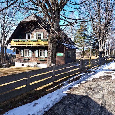 The Alm building with a balcony, green shutters, and a wooden fence along the road.