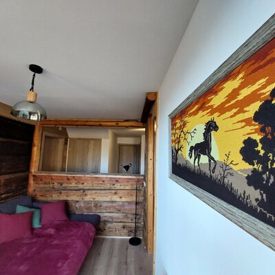 The Alpine Hut's living area features a sofa bed, wooden wall paneling, a framed horse tapestry, and an overhead pendant light.