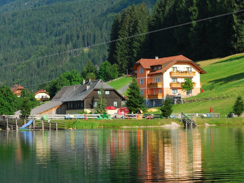 The Farm House property features a swimming lake with a pier and sun loungers, adjacent to the main aparthotel building and surrounding mountains.