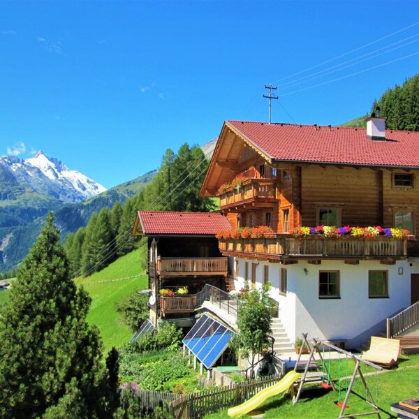 The farmhouse with wooden balconies, colorful flower boxes, a playground and trampoline in the garden, surrounded by the mountain landscape.