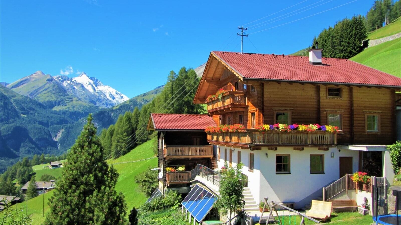 The farmhouse with wooden balconies, colorful flower boxes, a playground and trampoline in the garden, surrounded by the mountain landscape.