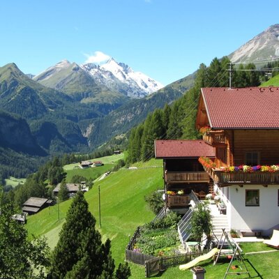 The farmhouse, featuring a wooden facade, red roofs, and balconies with flowers, is set amidst green mountains and offers a garden with a trampoline and slide.