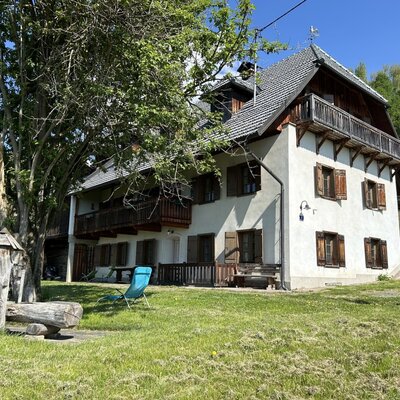 The vacation rental exterior features white walls, wooden balconies with shutters, and a garden with a large tree and a wooden water feature.