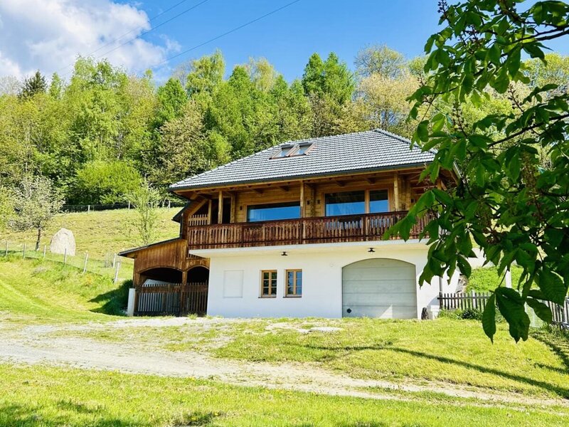 The vacation rental building, featuring a wooden upper level with a balcony and a white lower level with a garage, is surrounded by a grassy area and trees.