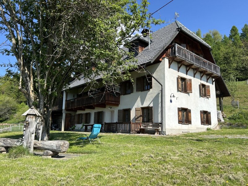The vacation rental exterior features white walls, wooden balconies with shutters, and a garden with a large tree and a wooden water feature.