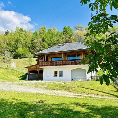 The vacation rental building, featuring a wooden upper level with a balcony and a white lower level with a garage, is surrounded by a grassy area and trees.