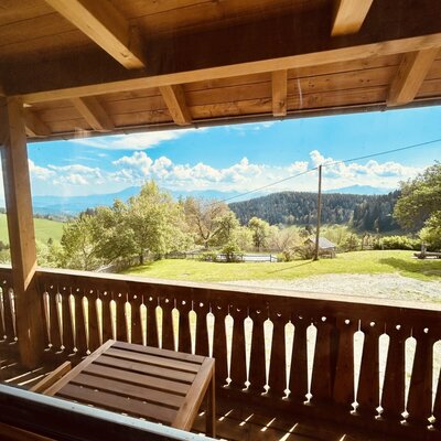 The covered wooden balcony of the vacation rental includes a small table and views of the hilly landscape and distant mountains.